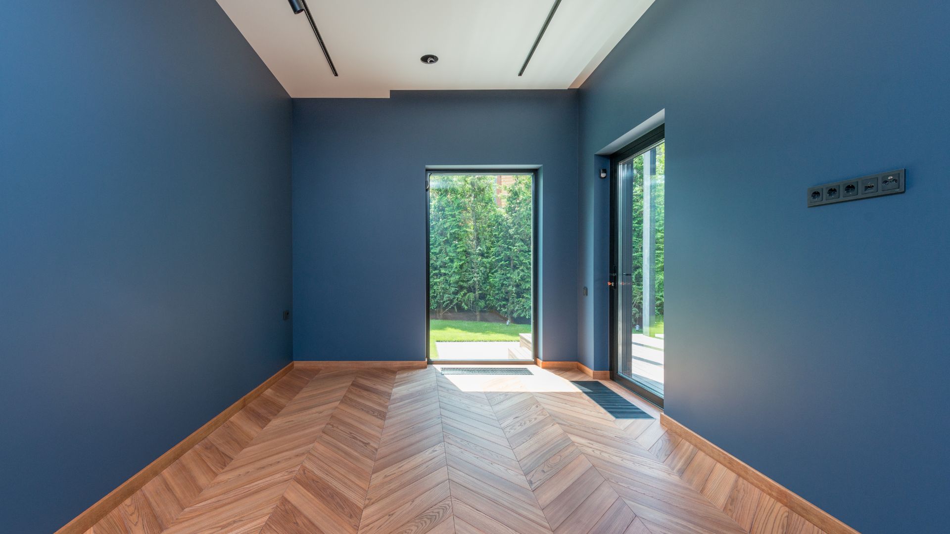 Empty blue room with herringbone wood floor and large windows to garden