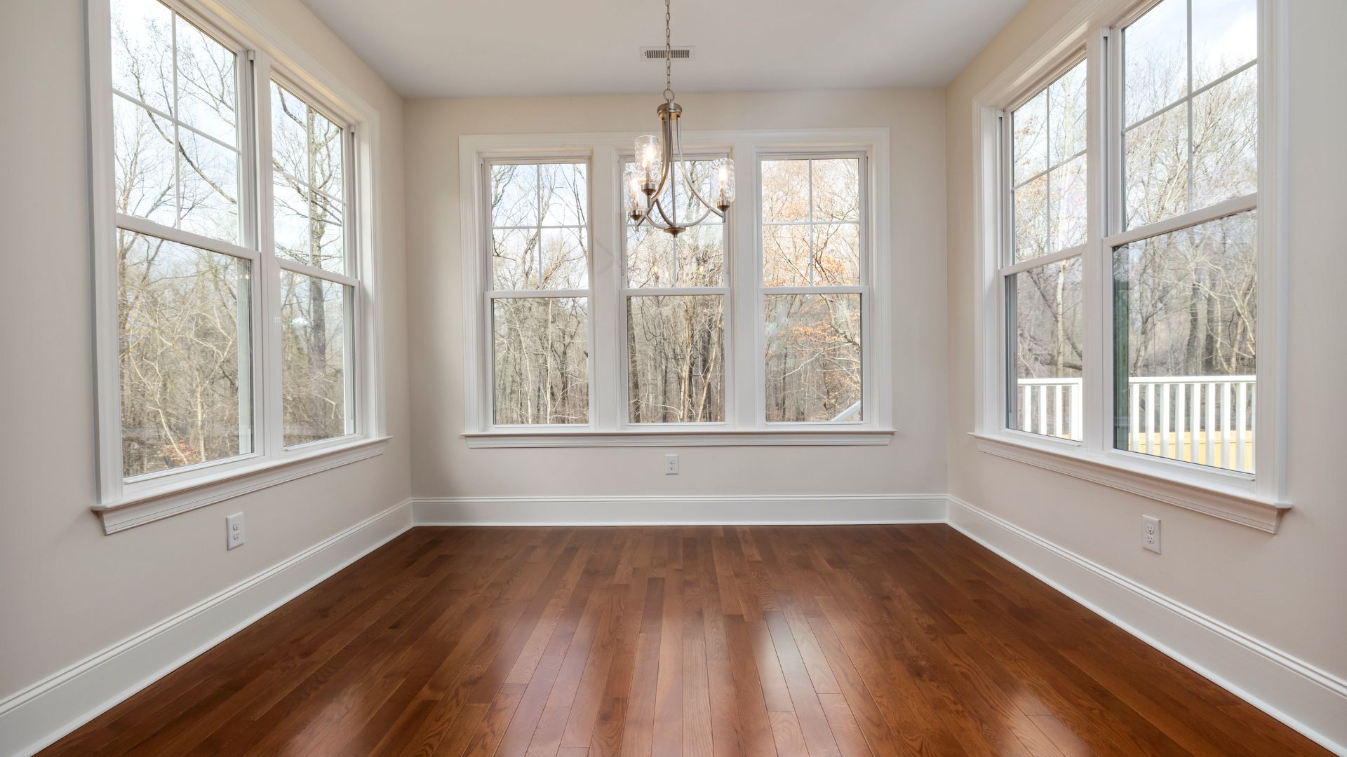 Empty room with large windows, hardwood floors, and chandelier overlooking forest