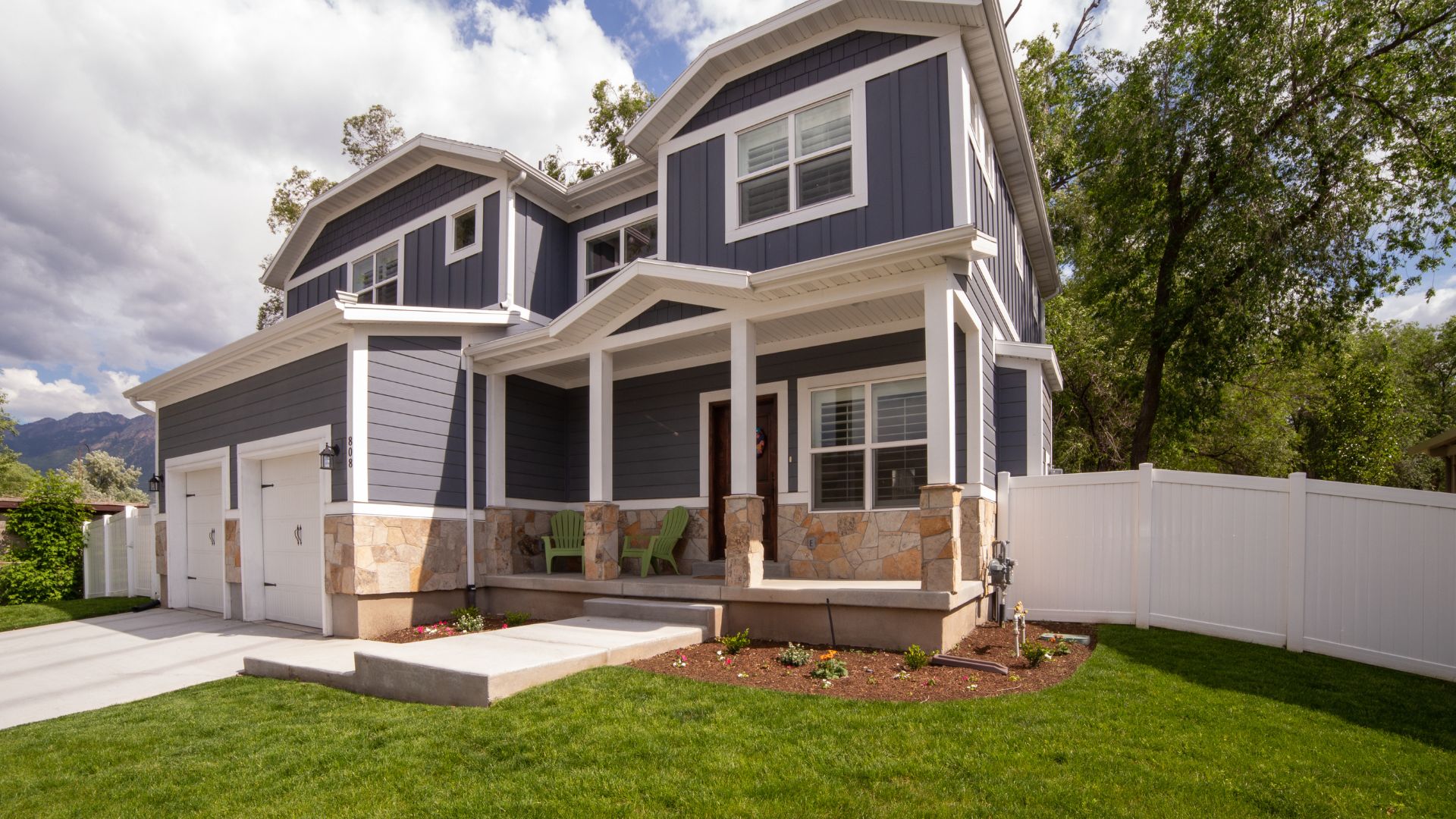 Two-story gray house with stone base, white trim, and green chairs on porch